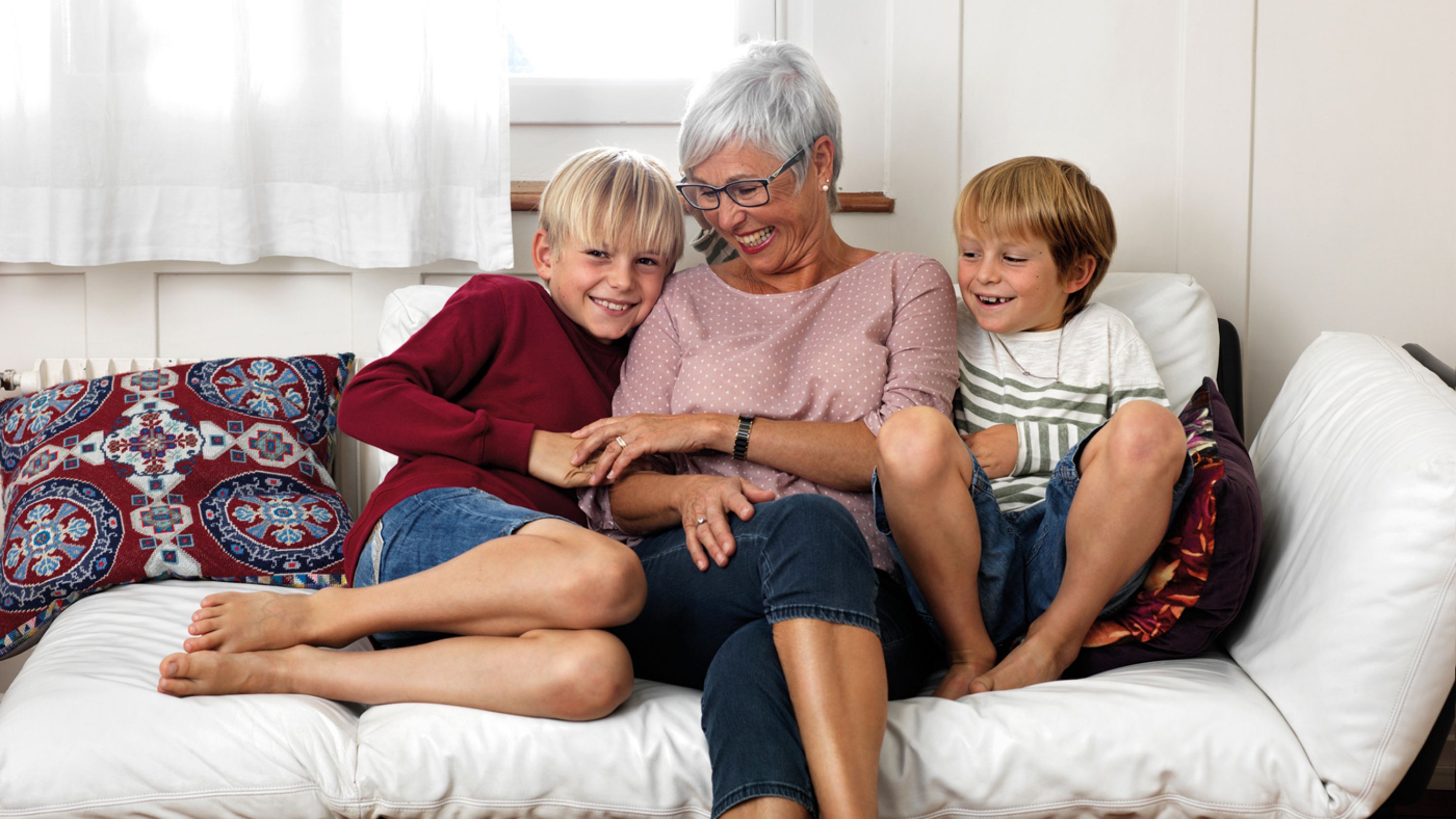 Eine Seniorin sitzt mit ihren zwei Enkelkindern auf einem Sofa und lachen